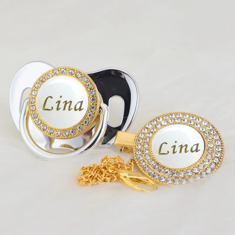 A gold-toned pacifier featuring a rhinestone name plate for Lina attached to a clip.