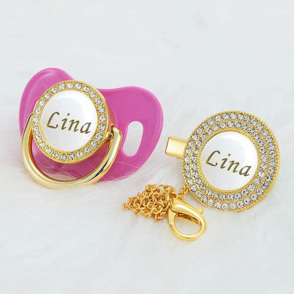 A pink custom name pacifier featuring a gold clip and a rhinestone-encrusted circular charm with the name Lina.