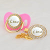 A gold-toned rhinestone pacifier featuring the name Lina on a white disc, attached to a pink clip.