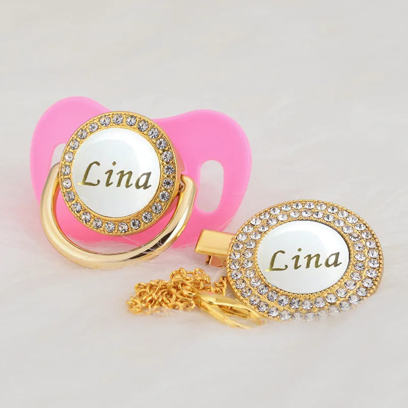 A gold-toned rhinestone pacifier featuring the name Lina on a white disc, attached to a pink clip.