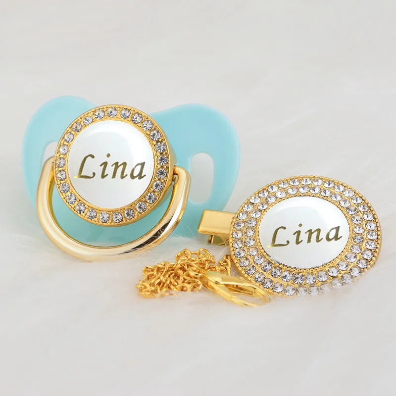 A custom name rhinestone pacifier featuring the name Lina set in a gold clip and light blue silicone.