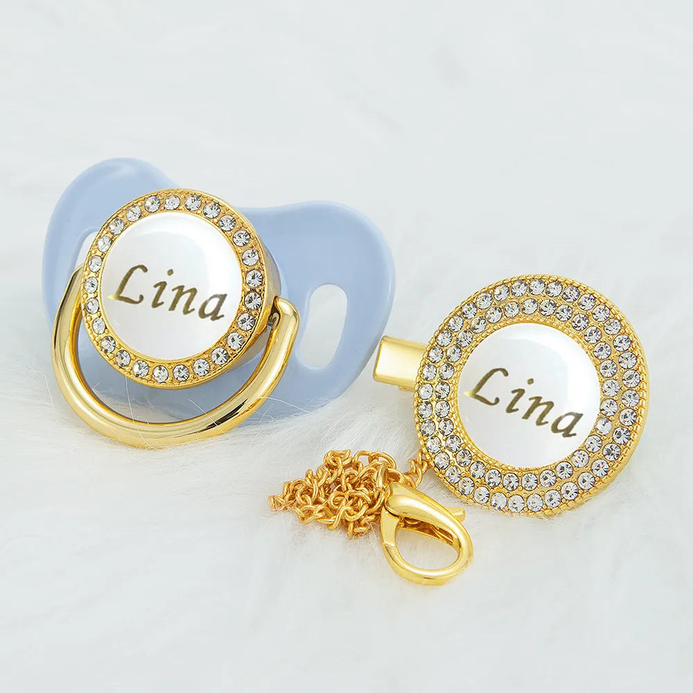 Gold-toned rhinestone pacifier with a white disc engraved with the name Lina and a light blue clip.