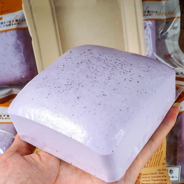 A hand holds a purple squishy stress relief toy shaped like a rectangular block with small black specks.