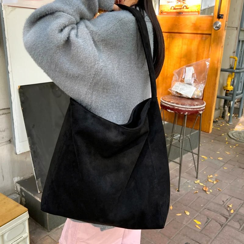 A person is holding a black suede tote shoulder bag with a gray sweater in an outdoor setting.