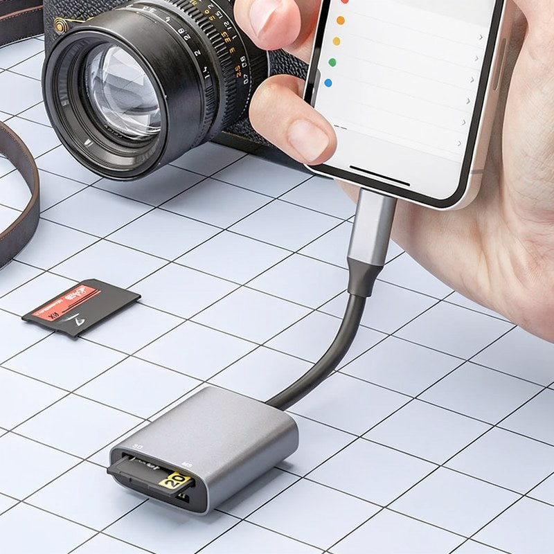 A hand holds a smartphone connected to a dual-slot card reader, which is positioned near a camera lens.