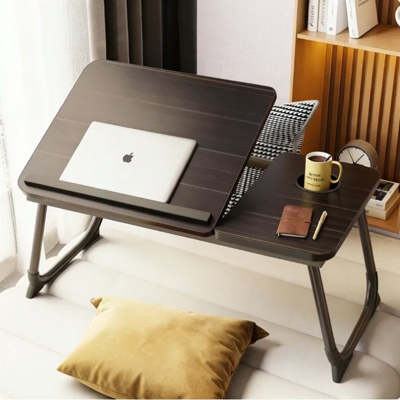 Un escritorio portátil de madera oscura junto a una cama con una almohada amarilla y un reloj pequeño.