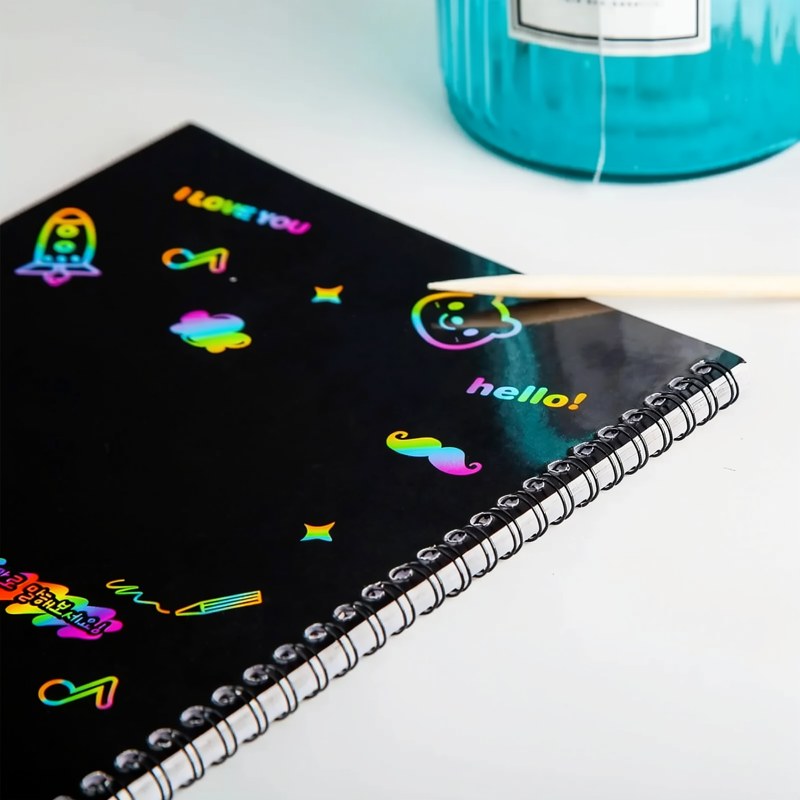 Un carnet spiral noir avec des dessins de scratch colorés, à côté d'un stylet en bois et d'un pot bleu sarcelle.