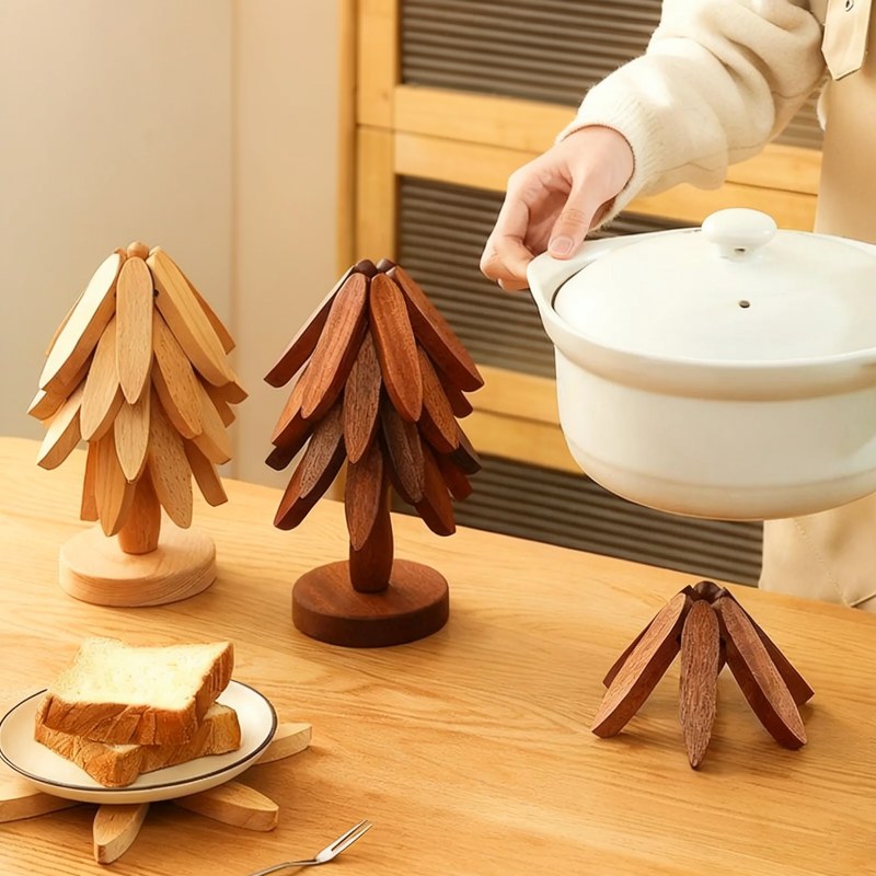Eine Hand hält einen weißen Topf mit Deckel über mehrere hölzerne, faltbare Untersetzer auf einem hellen Holztisch.