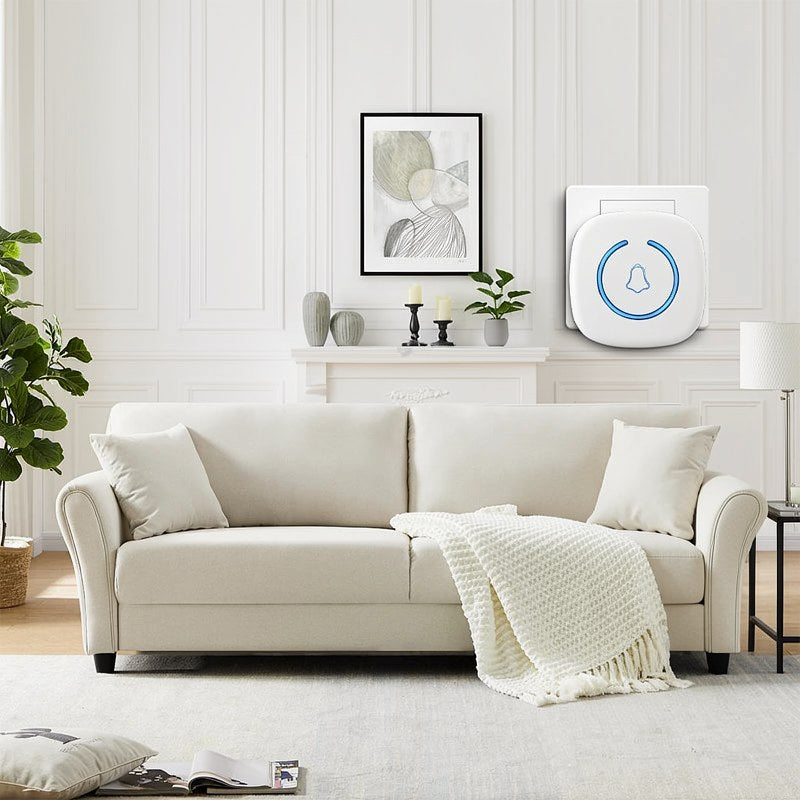 A white wireless doorbell is mounted on a wall above a sofa and artwork in a bright living room setting.