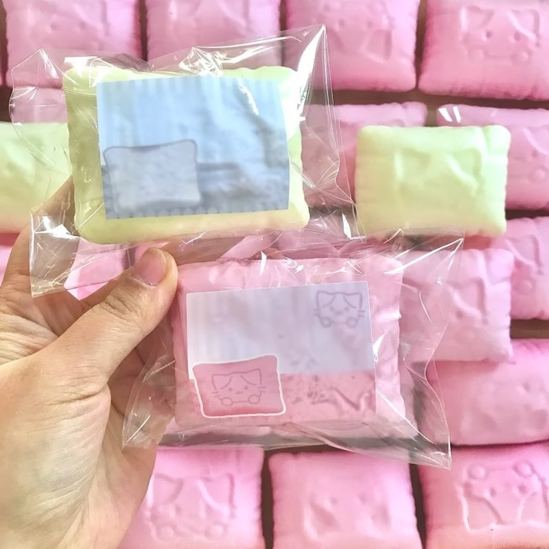 Une main tient un sac plastique transparent contenant un oreiller anti-stress en forme de chat, de couleur jaune pâle.