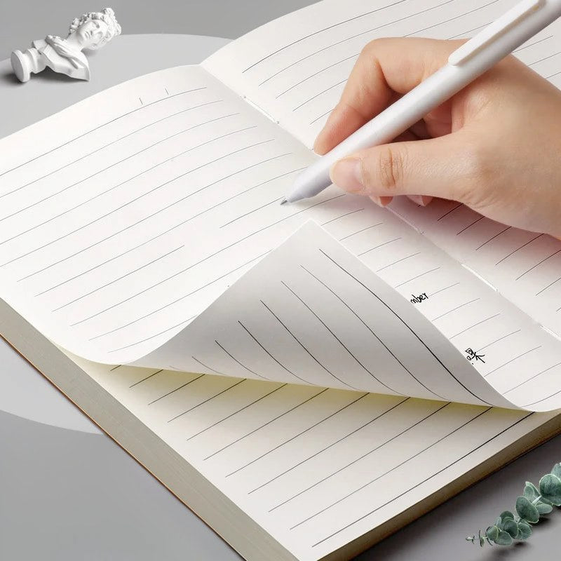Eine Hand schreibt in einem minimalistischen Kuhfell-Notizbuch mit karierten Seiten und einer kleinen weißen Skulptur daneben