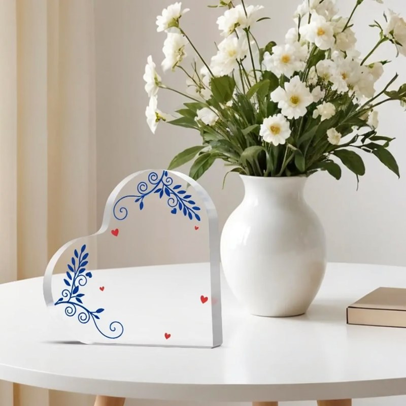 Weißes Acrylherzplakette mit blauen Blumenmustern auf weißem Tisch neben einer Vase mit weißen Blumen.