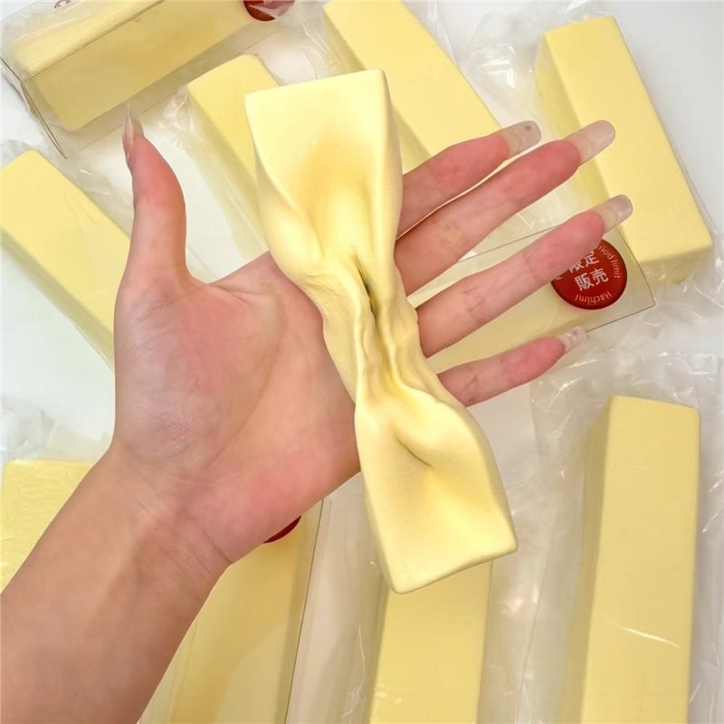 A hand is holding a yellow squishy butter block fidget toy with a satin ribbon tied around it.